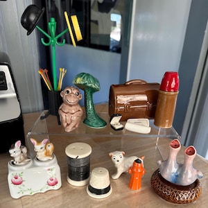 May include: A collection of ceramic figurines, including a monkey, a tree, a briefcase, and a lipstick. Other figurines include a salt and pepper shaker, a spool of thread, a thimble, a dog, a fire hydrant, and a snail. A green coat rack holds a black bowler hat.