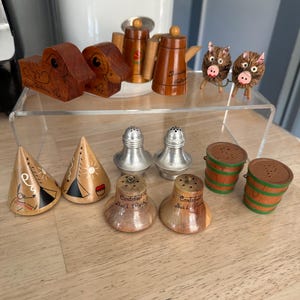 May include: A collection of vintage wooden salt and pepper shakers, including teapot, barrel, and cone-shaped designs. The set also features carved animal figurines and metal shakers. The items are displayed on a clear acrylic shelf.