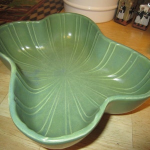 May include: A green ceramic serving bowl with a unique, leaf-like shape. The bowl has a smooth, glossy finish and features a series of raised lines that radiate from the center.