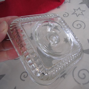 May include: A clear, square glass dish with a decorative rope-like border. The dish has a central, raised circular design with star-shaped details. The glass is held in a hand, with a red fabric visible in the background.