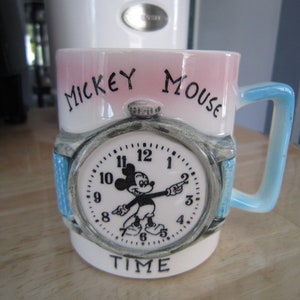 Puede incluir: Una taza de cer&aacute;mica blanca con un dise&ntilde;o rosa y azul. La taza presenta una esfera de reloj de Mickey Mouse con las palabras "Mickey Mouse Time" impresas en la parte delantera.