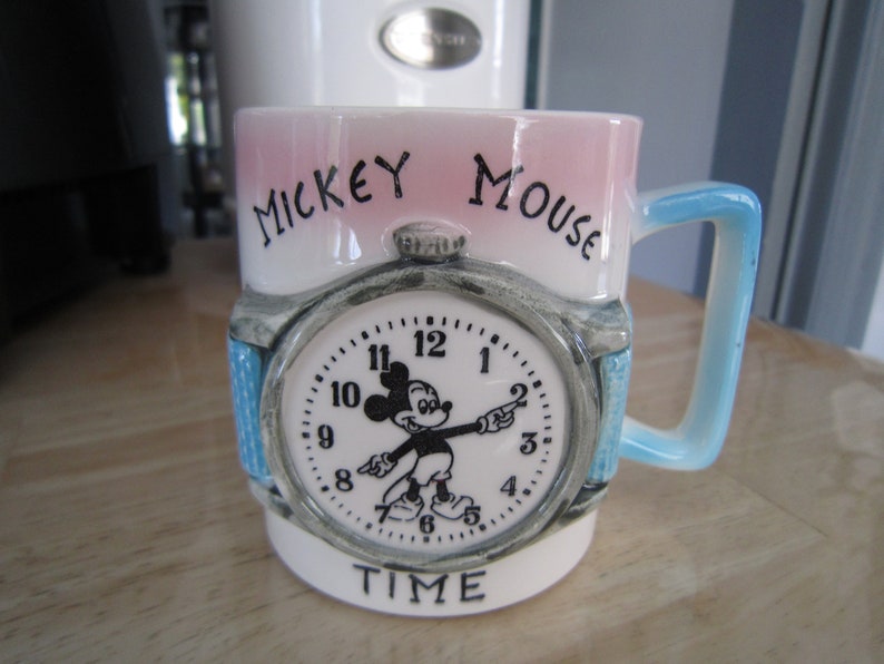Puede incluir: Una taza de cer&aacute;mica blanca con un dise&ntilde;o rosa y azul. La taza presenta una esfera de reloj de Mickey Mouse con las palabras "Mickey Mouse Time" impresas en la parte delantera.
