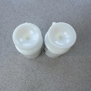 May include: Two white milk glass mugs viewed from directly above, showing the circular bases and the smooth, opaque finish of the glass. Each mug features a small handle extending from the side, with a subtle graphic visible on one side.