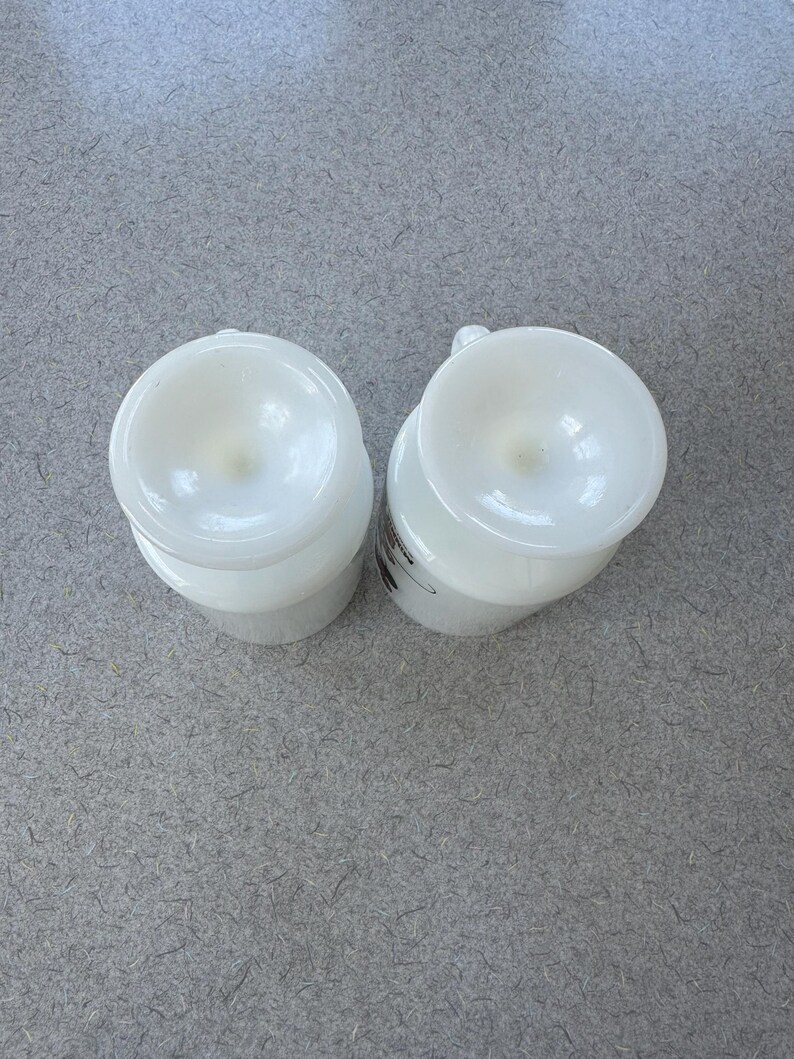 May include: Two white milk glass mugs viewed from directly above, showing the circular bases and the smooth, opaque finish of the glass. Each mug features a small handle extending from the side, with a subtle graphic visible on one side.