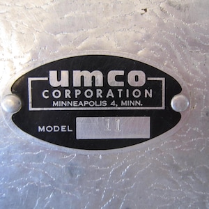 May include: A close-up of a metal plate with the text "umco corporation Minneapolis 4, Minn." in white lettering on a black oval. The plate also has the word "model" and the number "11". The plate is attached to a textured metal surface.