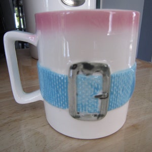 Puede incluir: Taza de cer&aacute;mica blanca con un dise&ntilde;o degradado rosa y blanco. La taza tiene un dise&ntilde;o de cintur&oacute;n azul y blanco con una hebilla plateada.