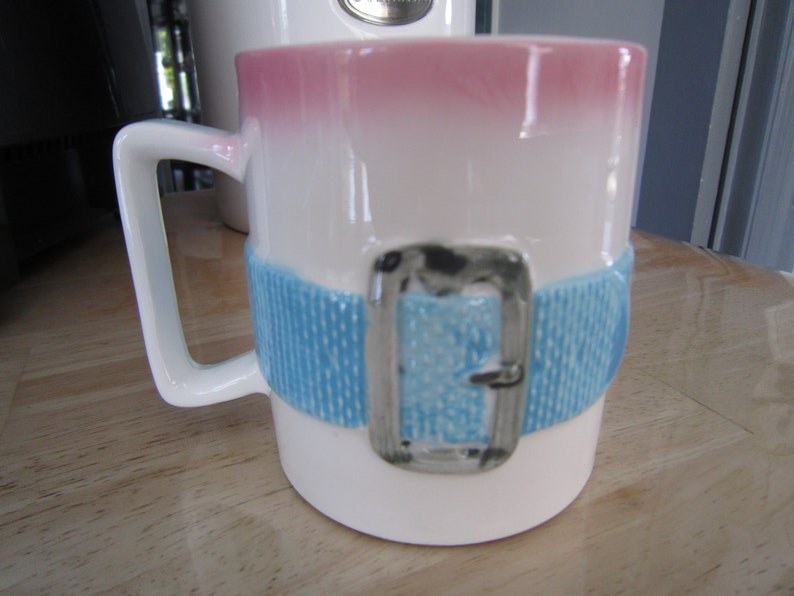 Puede incluir: Taza de cer&aacute;mica blanca con un dise&ntilde;o degradado rosa y blanco. La taza tiene un dise&ntilde;o de cintur&oacute;n azul y blanco con una hebilla plateada.