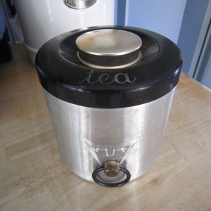 May include: A silver tea canister with a black lid and a silver knob. The lid has the word "tea" written on it in gold lettering.