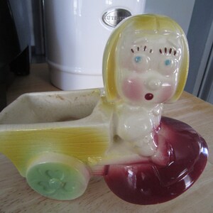 May include: A vintage ceramic planter shaped like a girl in a red dress pushing a yellow wagon. The girl has blonde hair, blue eyes, and a surprised expression.