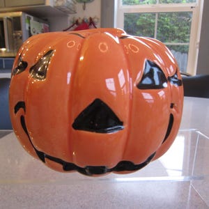 May include: A glossy orange ceramic jack-o'-lantern with a smooth, rounded pumpkin shape. It features carved-out triangular eyes and a nose, along with a wide, black-painted mouth. The surface has a reflective finish with deep vertical ridges.