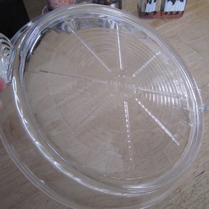 Puede incluir: Un molde para tartas de vidrio transparente con un borde acanalado y un asa a cada lado. El molde tiene un centro elevado con un dise&ntilde;o circular y las palabras "Fire King" impresas en la parte inferior.