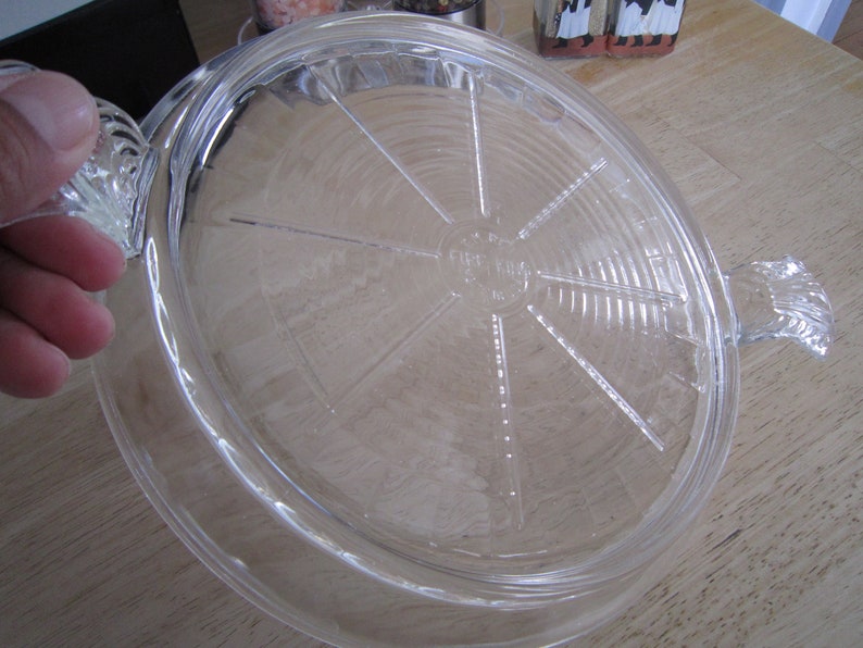 Puede incluir: Un molde para tartas de vidrio transparente con un borde acanalado y un asa a cada lado. El molde tiene un centro elevado con un dise&ntilde;o circular y las palabras "Fire King" impresas en la parte inferior.