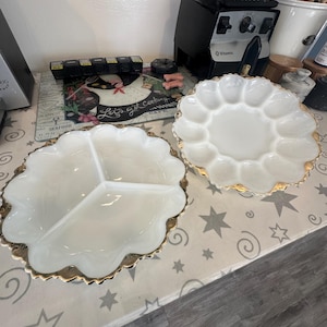 May include: Two white, scalloped-edge serving dishes with gold trim. One dish is divided into three sections, and the other has individual compartments. The dishes are on a patterned surface with a blender and other kitchen items in the background.