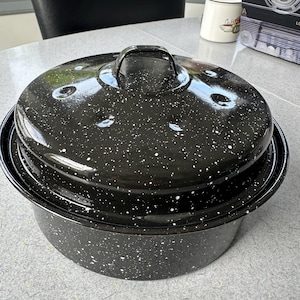 May include: A black enamel roasting pan with a lid. The pan and lid are speckled with white flecks. The lid has a handle for easy lifting. The pan is sitting on a gray surface.