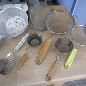 May include: A set of seven metal kitchen strainers with wooden and plastic handles. The strainers are of various sizes and shapes, including a colander, a fine mesh strainer, and a spoon strainer.