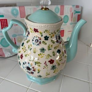 May include: A ceramic teapot featuring a cream-colored body adorned with a multi-colored floral pattern. The teapot includes a light blue lid with a clear glass knob, a matching light blue handle, and a matching light blue spout. The base is light blue.
