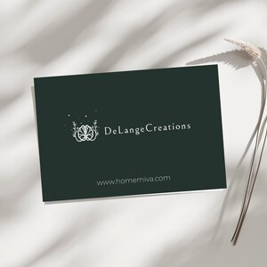 Op de afbeelding: Een donkergroene visitekaart met de witte tekst "DeLange Creations", een logo en het websiteadres www.homemiva.com. De kaart ligt op een witte achtergrond met schaduwen en een takje gedroogde bloemen.