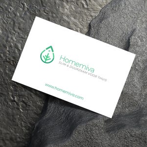 Op de afbeelding: Een witte visitekaartje met het logo en de tekst "Homemiva" in groen. Het logo is een groen blad en een huisontwerp. Het websiteadres www.homemiva.com wordt ook weergegeven. De kaart ligt op een gestructureerd, donkergrijs oppervlak.