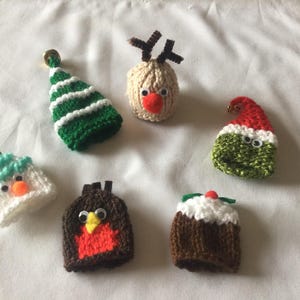 May include: Six knitted Christmas decorations, including a green and white Christmas tree, a reindeer, a Santa hat, a snowman, a robin, and a Christmas pudding. Each ornament is handmade.
