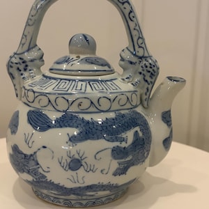 May include: A white ceramic teapot featuring blue hand-painted dragon motifs, cloud patterns, and geometric borders. The teapot has a high arched handle with sculpted dragon heads at the base, a matching lid with a knob, and a curved spout.