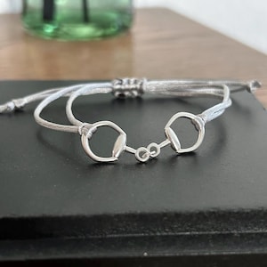 Könnte beinhalten: Ein graues geflochtenes Armband mit zwei silbernen Gebiss-Anhängern, die durch einen silbernen Ring verbunden sind.