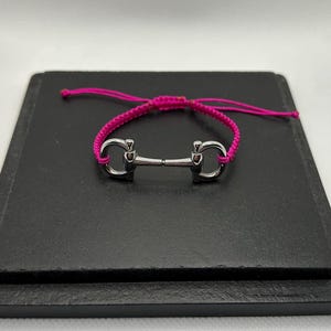 以下が含まれることがあります： 明るいピンク色の編み紐が付いた、銀色の馬具ビットブレスレット。ブレスレットは黒い正方形の表面に表示されています。ビットはクラシックなデザインで、紐はサイズ調整のために結ばれています。