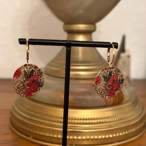 Könnte beinhalten: Ein Paar goldene Creolen mit runden Anhängern in Rot und Grün mit floralem Muster. Die Ohrringe sind auf einem schwarzen Ständer ausgestellt.