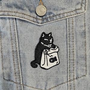 May include: A black cat with a white bag in its paws. The cat is sitting on a light blue denim jacket with a pocket. The cat is looking to the right.