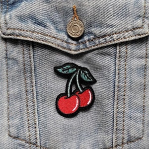 May include: A denim jacket with a pocket featuring a red cherry embroidered patch.