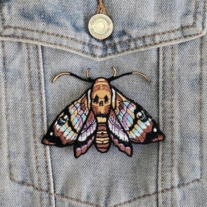May include: A light blue denim jacket with a pocket featuring an embroidered patch of a moth with a skull design. The moth has brown, purple, and pink wings.