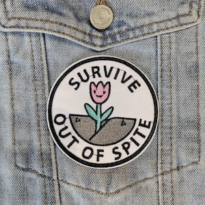 Pode incluir: Um remendo bordado branco e preto com uma flor rosa e o texto "Survive Out of Spite" em uma jaqueta jeans azul claro.