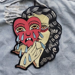 Puede incluir: Un parche bordado negro y rojo de una mujer sosteniendo un corazón con una cara llorando. El corazón es rojo con ojos amarillos y lágrimas azules. La mujer tiene el pelo negro y lleva un vestido amarillo. El parche está en una chaqueta vaquera azul claro.
