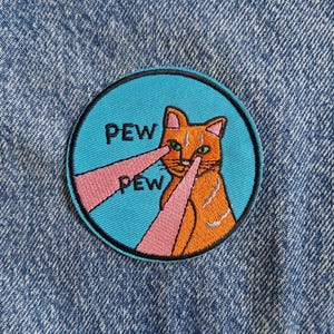 Op de afbeelding: Een ronde geborduurde patch met een oranje kat op een felblauwe achtergrond. De kat heeft groene ogen en schiet roze laserstralen uit de ogen. De tekst PEW PEW is in zwarte letters naast de laserstralen geborduurd.