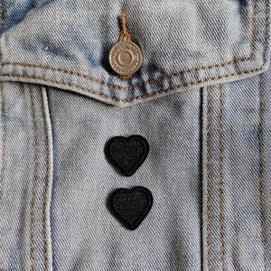 以下が含まれることがあります： ポケットとシルバーのボタンが付いた、明るいブルーのデニムジャケットのクローズアップ。2つの黒いハート型のパッチがジャケットに縫い付けられています。ジャケットには、コントラストの効いた茶色のステッチが見えます。