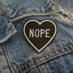 Peut inclure: Un patch en forme de cœur noir avec des coutures blanches et le mot "NOPE" en lettres blanches.