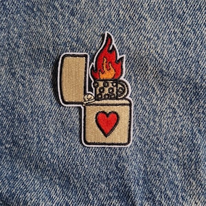 Puede incluir: Un parche bordado que muestra un encendedor beige con la tapa abierta. El encendedor tiene un corazón rojo en la parte frontal y una llama estilizada de color naranja y rojo que sale de la parte superior. El parche está sobre tela vaquera azul.