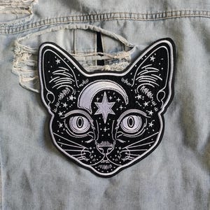 Puede incluir: Un parche bordado en blanco y negro con un gato, una luna creciente y una estrella. El parche está adherido a una chaqueta vaquera azul claro. La cara del gato presenta ojos grandes y detalles intrincados.