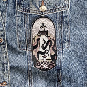 Peut inclure: Veste en jean avec un écusson représentant un phare. L'écusson a une bordure noire et représente un phare avec des tentacules noirs tourbillonnants et un ciel rose et gris. La veste a des boutons métalliques et des poches.