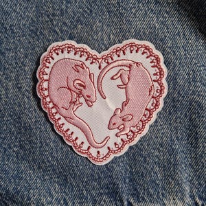 May include: A heart-shaped embroidered patch featuring two light pink rats positioned to form a heart outline. The design is enclosed by a decorative red scalloped border with small circular details, set against a blue denim fabric background.