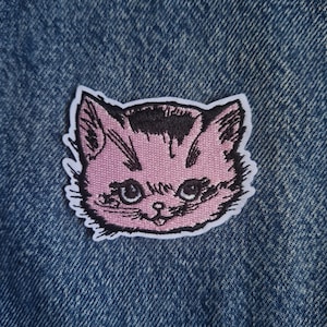 Puede incluir: Un parche bordado que muestra una cabeza de gato estilizada con pelaje rosa claro, sombreado rosa oscuro y detalles de líneas negras. El gato tiene ojos grandes, una nariz pequeña y un borde blanco, sobre un fondo de tela vaquera azul texturizada.