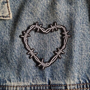 May include: A blue denim jacket featuring an embroidered patch in the shape of a heart made from barbed wire. The patch has a black outline with white and grey details, stitched onto the textured, medium-wash blue fabric of the garment.