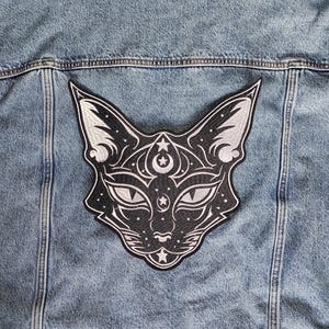 May include: A light blue denim jacket with a large, black and white cat patch on the back. The patch features a stylized cat face with celestial details like stars and a crescent moon.