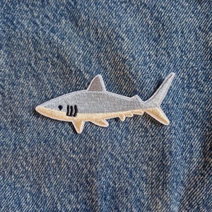 May include: An embroidered shark patch featuring a grey upper body and a light tan underbelly, attached to a blue denim fabric. The shark is depicted in profile with a small black eye and three black gill slits on its side, showing detailed stitching.