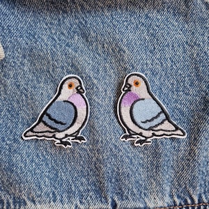 Könnte beinhalten: Zwei gestickte Aufbügler mit Taubenmotiv. Jeder Vogel hat hellgraue Federn, einen blauen Flügelbereich mit dunklem Streifen, einen violetten Halsfleck, ein orangefarbenes Auge und einen schwarzen Schnabel, alles mit weißem Garn auf Jeansstoff.