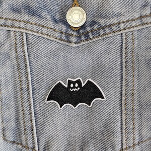 May include: A black bat with a white smiling face is ironed onto a blue denim jacket. The bat is centred on the jacket's pocket.