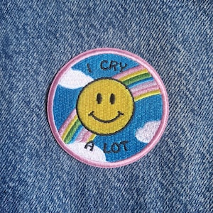 May include: A circular embroidered patch featuring a yellow smiley face in the centre against a blue sky background with white clouds and a rainbow. The text "I CRY A LOT" is stitched in black letters around the smiley face, bordered by pink.