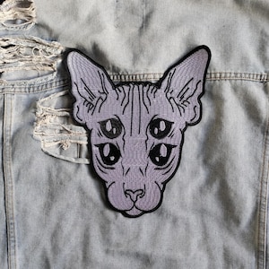 May include: A light blue denim jacket with distressed detailing. A grey patch of a four-eyed cat is sewn onto the back of the jacket. The cat patch has a black outline and detailed facial features.