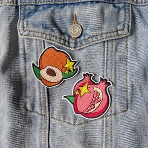 May include: Two embroidered patches attached to a light blue denim jacket. One patch depicts an orange peach with a brown pit and green leaves, and the other shows a pink pomegranate with visible seeds and green leaves. Both feature yellow stars.