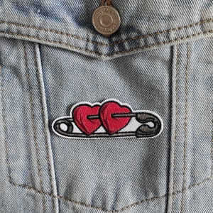 May include: A red and white embroidered patch of two hearts connected by a safety pin on a blue denim jacket.