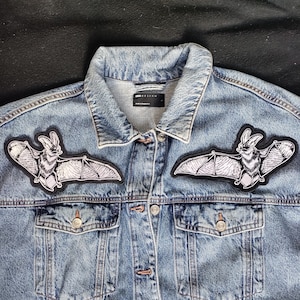 May include: Pale blue denim jacket featuring two bat-shaped patches on the chest. The patches are black and white, depicting detailed bat wings. The jacket has a button-up front, two chest pockets, and a classic collar.